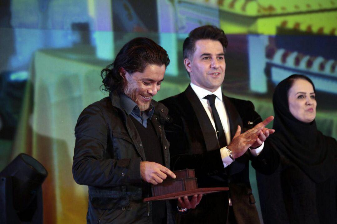 arash bahmani winning awards on stage آرش بهمنی برنده جایزه انجمنهای خیریه - arash bahmani with a black coat smiling and holds his award for the best film director on stage